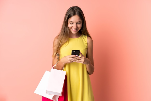 Young Lithuanian woman on pink holding shopping bags and writing a message with her cell phone to a friend