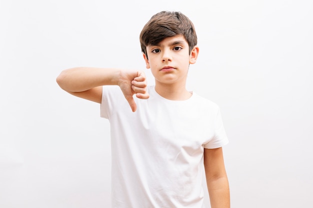 Молодой маленький мальчик в белой футболке, стоящий на белом изолированном фоне, выглядит несчастным и сердитым, показывая отказ и отрицательный жест пальцами вниз. Плохое выражение.