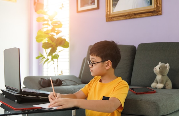 Young little boy wearing eyesglasses sitting near sofa in living room at home and do homework during class learning via laptop notebook computer.