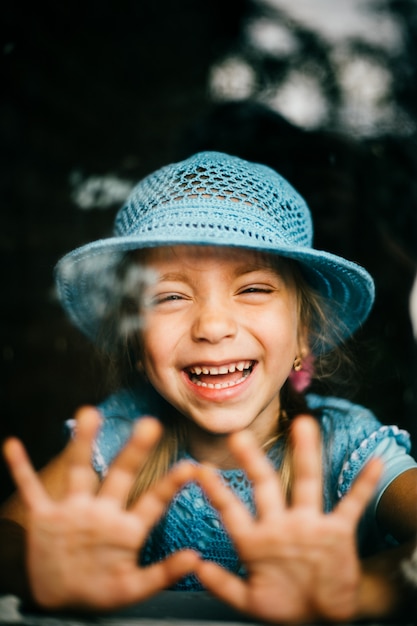 Young little girl making faces and laughing through window with palms lying on it. Expressive child emotions. Dramatic happy kid face.