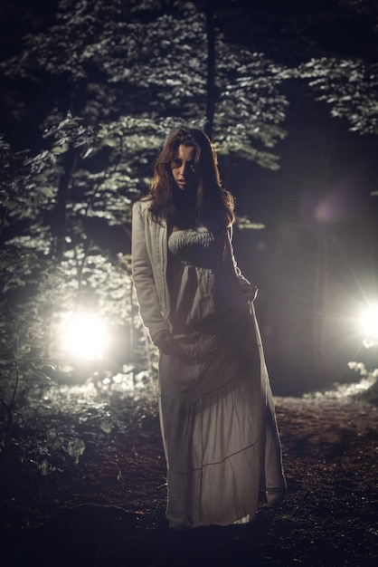 Young lonely woman walking through the forest at night in white dress. Looking at camera.