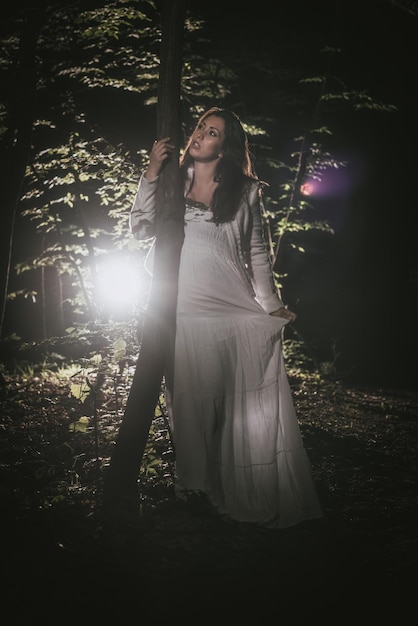 Young lonely woman walking through the forest at night in white dress.