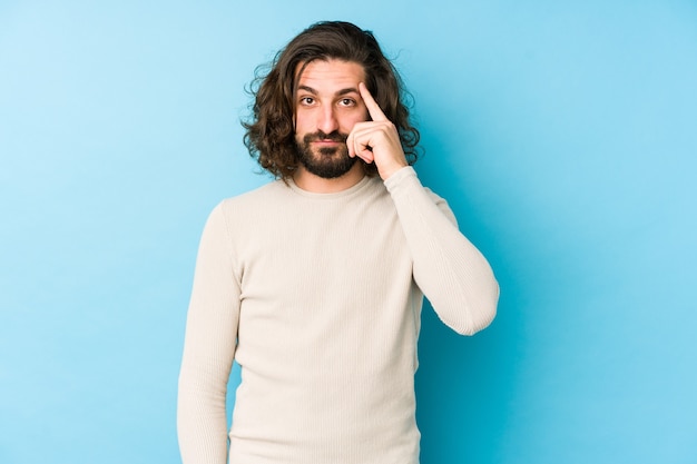 Young long hair man isolated on a blue wall pointing temple with finger, thinking, focused on a task.