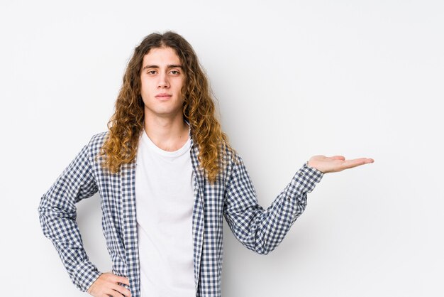 Young long hair man posing isolated showing a copy space on a palm and holding another hand on waist.