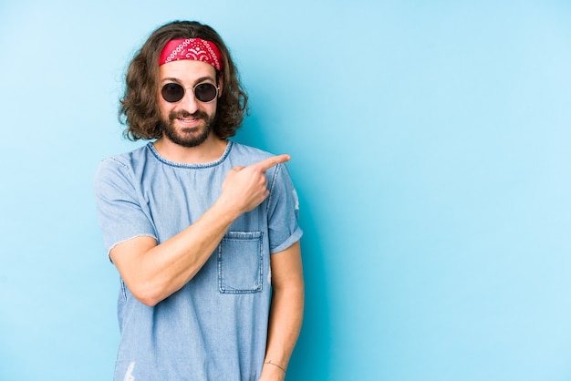 Young long hair man wearing a festival hipster look isolated smiling and pointing aside, showing something at blank space