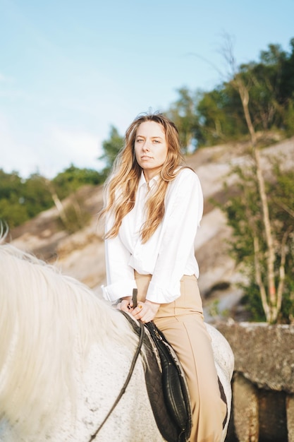 Foto giovane donna dai capelli lunghi in camicia bianca a cavallo bianco sullo sfondo del paesaggio marino