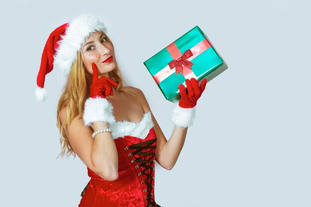 Young long-haired blonde in a Santa Claus dress, holding a gift in her hand and looking straight ahead.