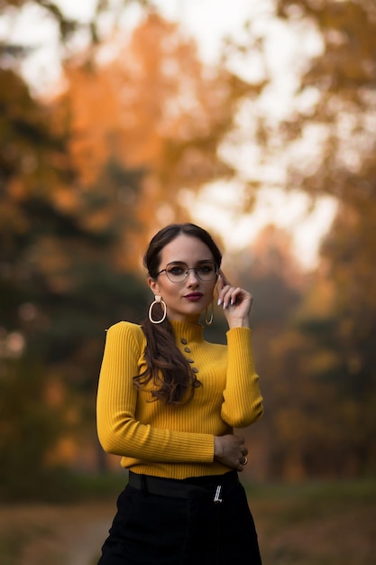 Young long haired brunette in yellow knitted jumper and eyeglasses looking at camera while standing against blurred foliage of autumn trees in park