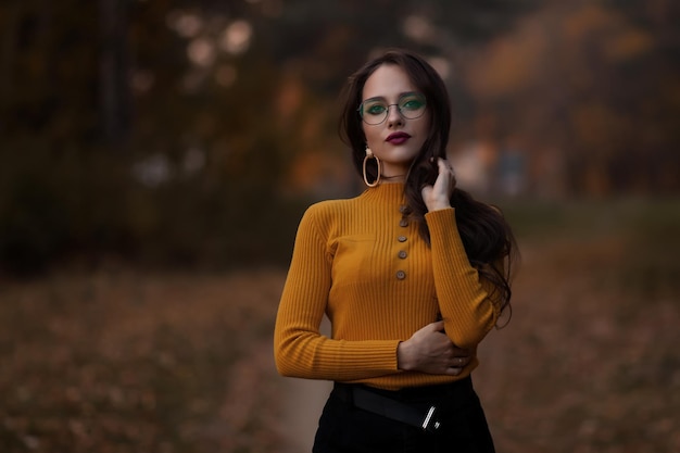 Young long haired brunette in yellow knitted jumper and eyeglasses looking at camera while standing against blurred foliage of autumn trees in park