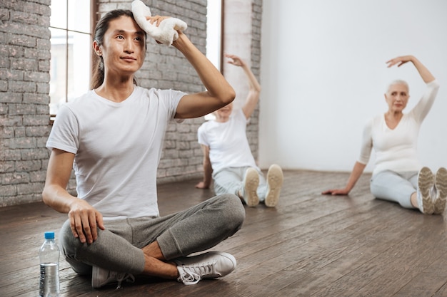 Young longhaired instructor wearing white T-shirt holding his back straight while crossing both legs