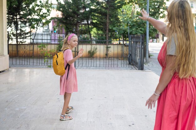 Young longhaired mother and little schoolgirl wave goodbye before classes near school