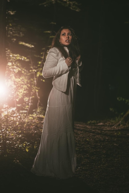 흰 드레스를 입고 밤에 숲을 걷고 있는 젊은 잃어버린 여자.