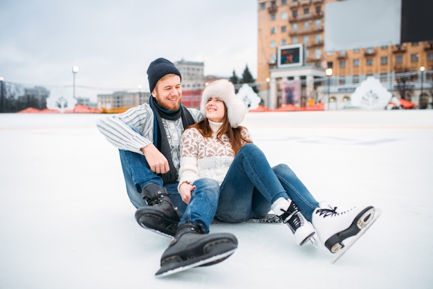 Young love couple in skates sitting on ice, skating rink. Winter ice-skating on open air, active leisure