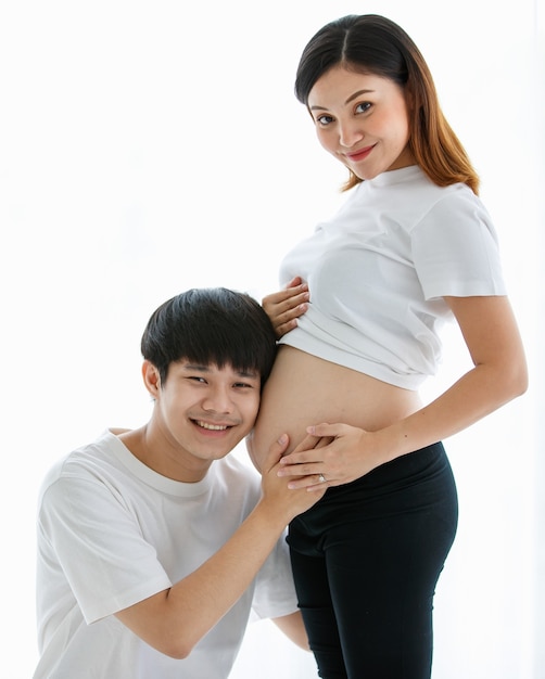 Молодые милые азиатские муж и жена остаются вместе в гостиной. Беременная женщина стоит и смотрит на свой живот, в то время как мужчина сидит и слушает ребенка в ее животе с любовью, копируя пространство