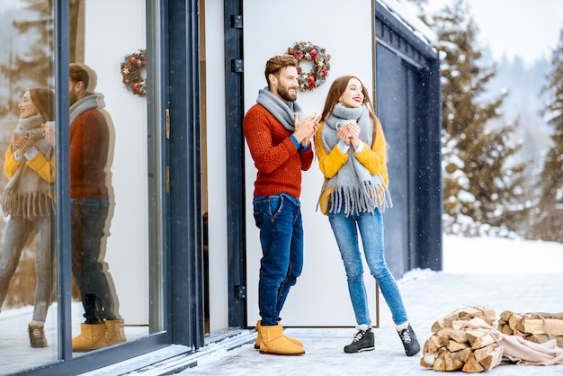 Young lovely couple dressed in colorful sweaters enjoying beautiful view standing with hot drinks on the terrace of the modern house in the mountains