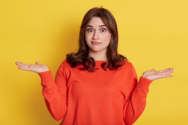 Young lovely female with dark hair, having clueless and unaware expression, purses lips, puzzled, isolated over yellow wall.