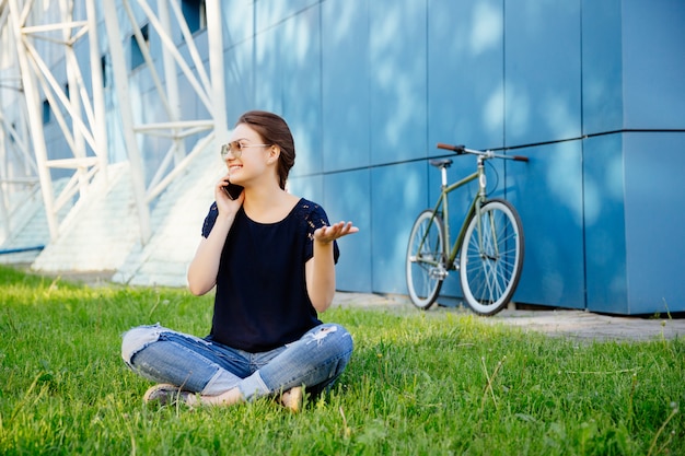 自転車で歩いた後、草の上に座って、携帯電話で話し、余暇を楽しんでいるカジュアルな服装の若い素敵な女性。