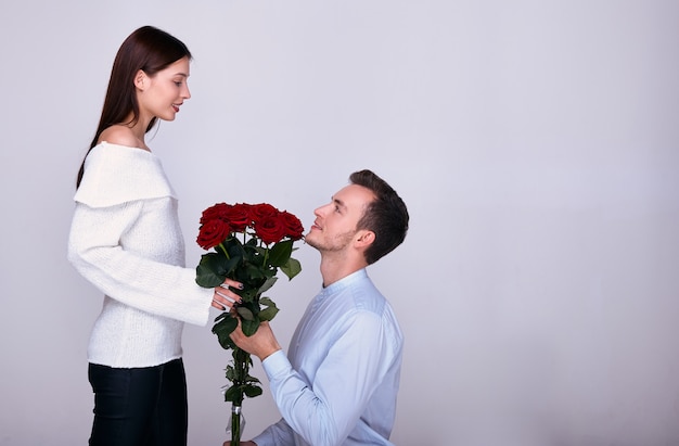A young lover stands on one knee and gives his womanfriend red roses.