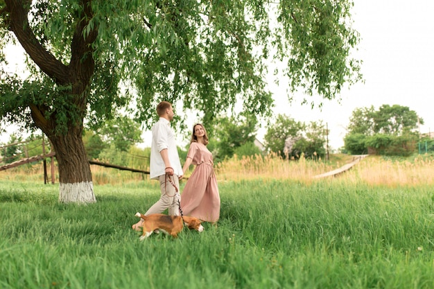 Young loving couple having fun and running on the green grass on the lawn with their beloved domestic dog breed Beagle and a bouquet of wildflowers