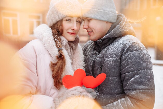 Young loving couple outside on a winter day holding red paper hearts in their hands feeling joy and love. Romantic man and woman celebrate Valentine's day under the snow at a park. 