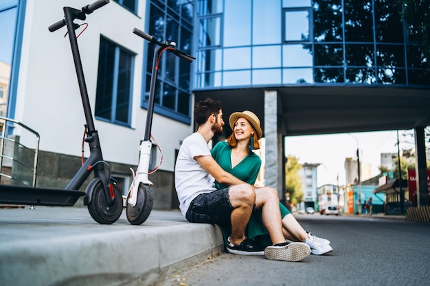 Young loving couple relaxing sitting near a modern glass building with their electric scooters. Walks in the big city