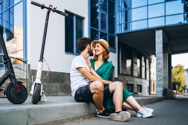 Young loving couple relaxing sitting near a modern glass building with their electric scooters.