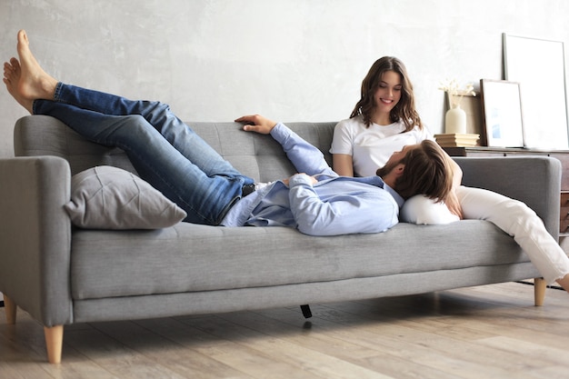 Young loving couple relaxing on sofa together, husband lying on wife legs resting on couch, concept of capture happy moment.