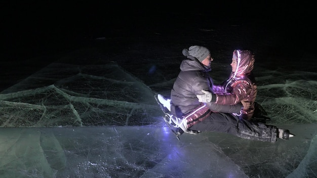 Young loving couple skating at ice rink at night Man and woman kiss hug have fun learn to skate Making love on ice Beautiful clear ice with cracks on lake Ice illuminated backlit from below