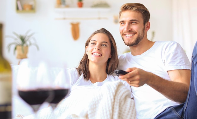 Giovane coppia di innamorati sul divano di casa a guardare la tv e ridere bevendo un bicchiere di vino rosso