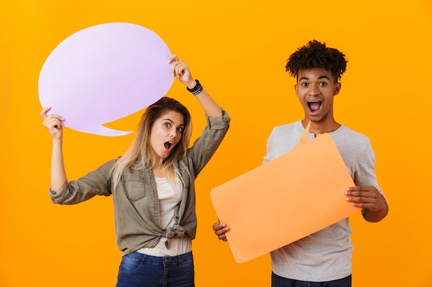 Young loving couple standing isolated over yellow wall holding speech bubble