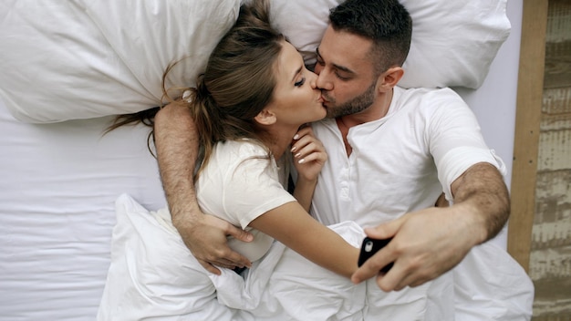 Young and loving couple take selfie picture on smartphone camera and kiss while lying in bed