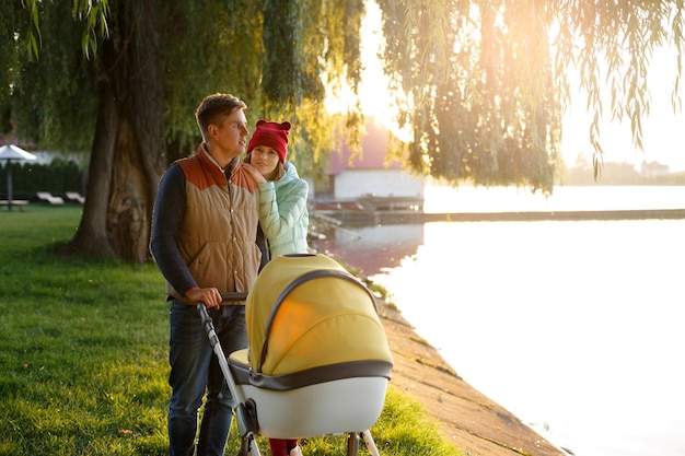 A young loving family walks by the lake with a stroller smiling parents couple with baby pram in