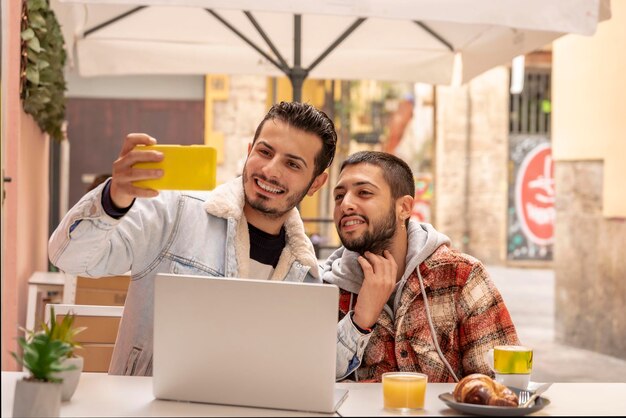 Молодая любящая гей-пара делает селфи, используя ноутбук и завтракая в кафе