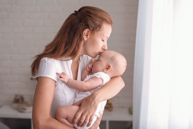 Young loving mother hugs and kisses her baby at the bedroom window.