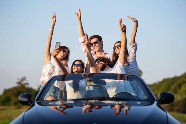 Giovani ragazze e ragazzi fortunati in occhiali da sole sono seduti in una cabriolet nera sulla strada alzando le mani e facendo selfie in una giornata di sole. .