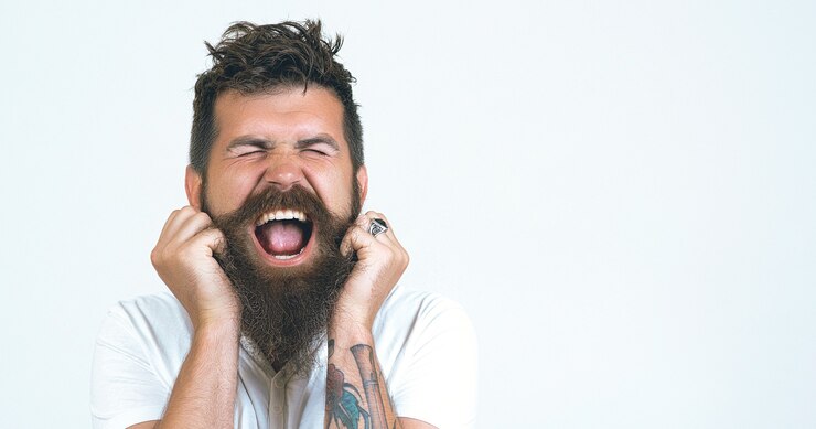Photo young mad man, expressive face, scared pos. portrait stressed and screaming crazy handsome bearded man wearing white t-shirt on white wall background. nerves at the limit when the beard is scratched