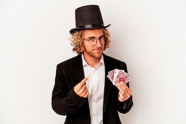 Young magician caucasian man holding magic cards isolated on white background pointing with finger at you as if inviting come closer.