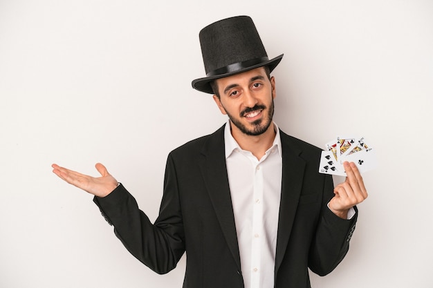 Young magician man holding a magic card isolated on white background showing a copy space on a palm and holding another hand on waist.