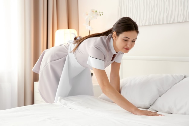 Young maid making bed in light hotel room