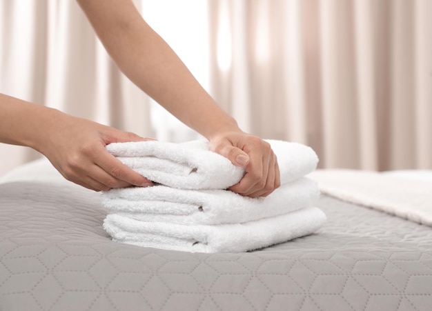 Young maid putting stack of fresh towels on bed in hotel room closeup