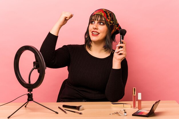 Young makeup artist woman doing a makeup tutorial isolated on pink background raising fist after a victory winner concept