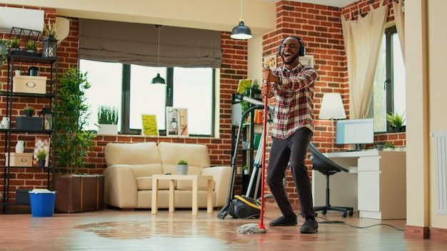 Giovane maschio adulto che si diverte con mop e musica a casa, mostrando passi di danza e cantando in soggiorno. persona felice che pulisce i pavimenti in legno e fa le pulizie di primavera, i lavori domestici.