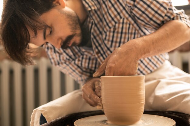 Young male artisan working over new clay item while shaping it during rotation in pottery wheel