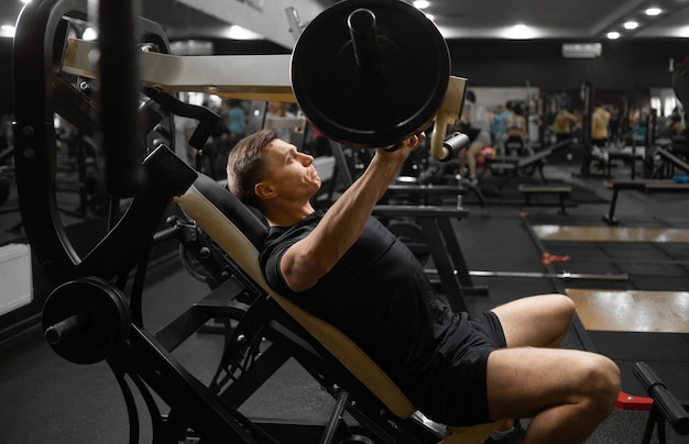 A young male athlete on the simulator in a sitting position
shakes the muscles of his arms and chest athlete side view