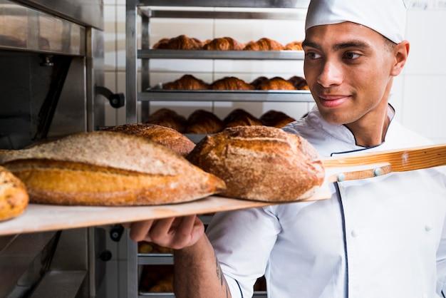 Photo young male baker taking out with wooden shovel freshly baked bread from the oven