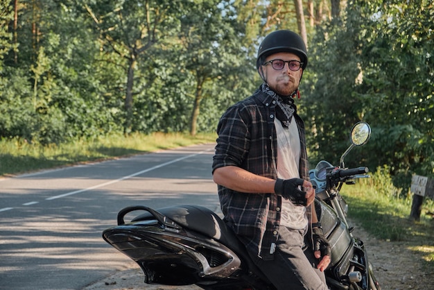 Young male biker in helmet travels on a motorcycle alone stopped and smoking on the side of a forest