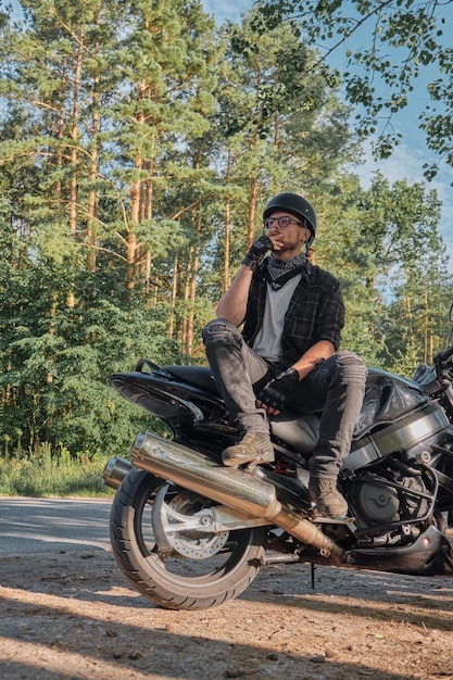 Young male biker in helmet travels on a motorcycle alone stopped and smoking on the side of a forest