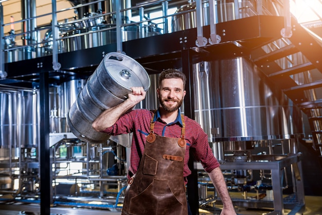 Young male brewer in leather apron holds barrel with craft beer at modern brewery factory