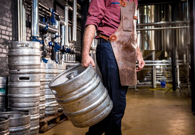 Young male brewer in leather apron holds barrel with craft beer at modern brewery factory