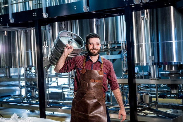 Young male brewer in leather apron holds barrel with craft beer at modern brewery factory